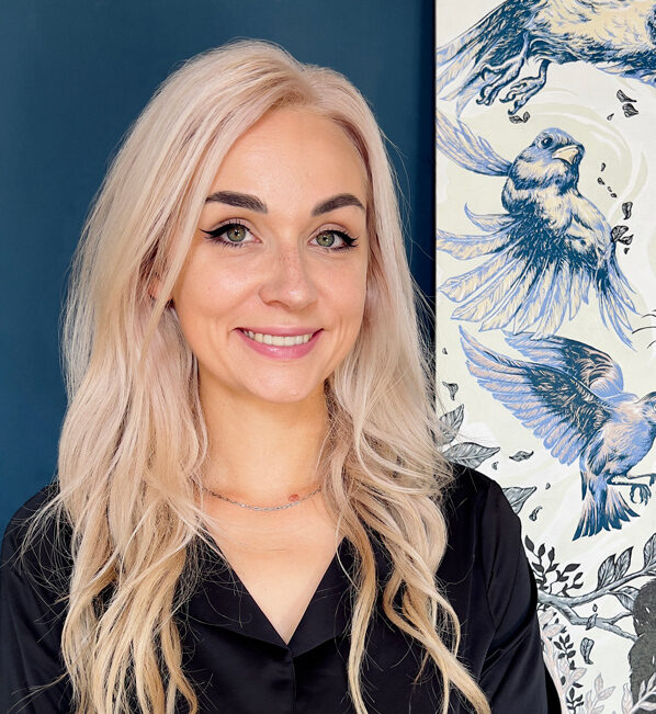 A smiling woman with long, wavy, light pink hair and a black shirt stands in front of a dark blue wall and a painting of blue birds.