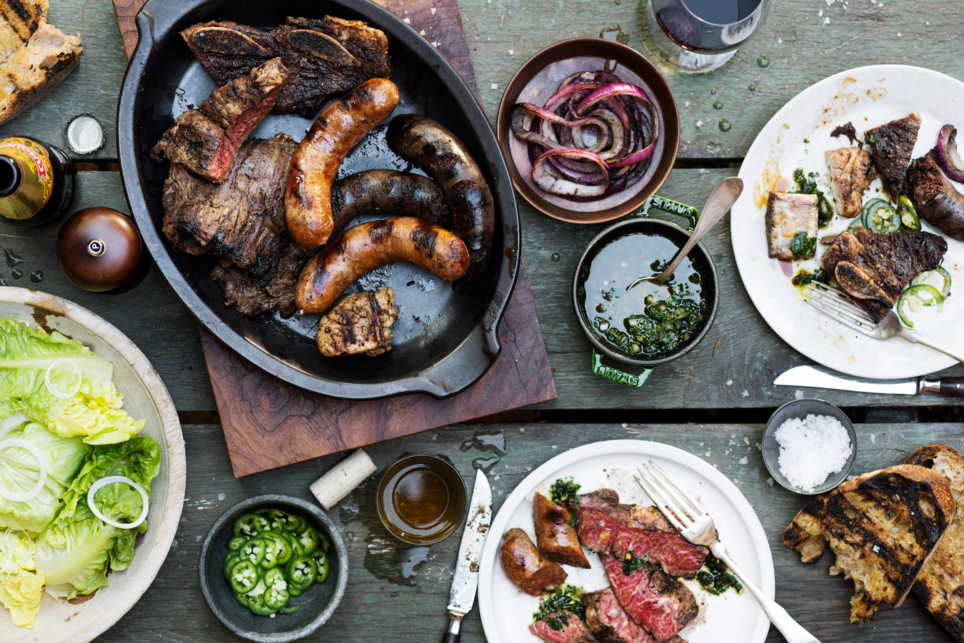 Blick von oben auf gegrilltes Fleisch, Würstchen, Chimichurri-Sauce und Beilagen auf einem rustikalen Tisch. Foto von Evan Sung