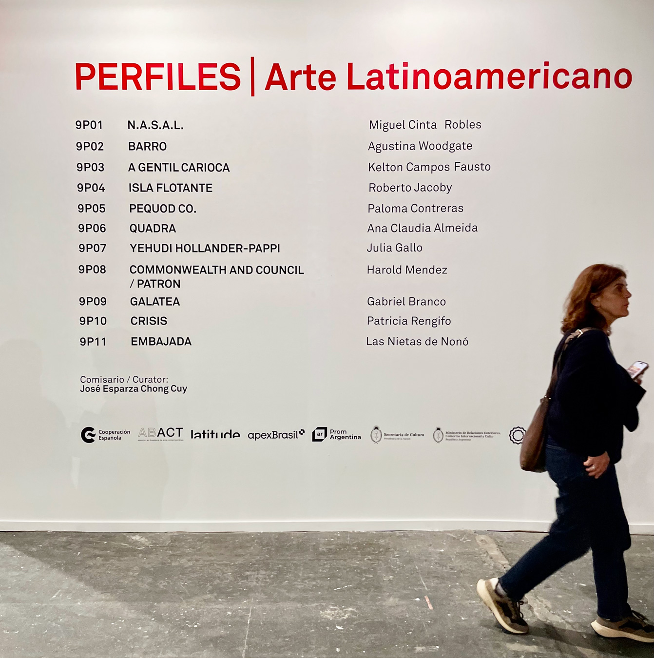 Una mujer pasa junto a una pared blanca con el título "PERFILES | Arte Latinoamericano" y una lista de nombres y organizaciones en texto negro. En la pared también aparecen logotipos de varios patrocinadores y colaboradores de la feria de arte ARCOmadrid 2026.