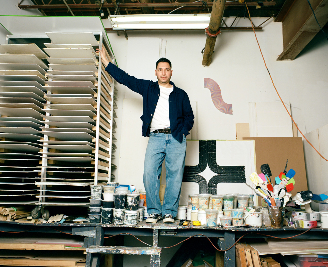 Der Künstler Robert Davidovitz steht in seinem Atelier, gelehnt an ein Regal mit trocknenden Leinwänden, umgeben von Farbdosen, Pinseln und Kunstwerken. Er trägt eine marineblaue Jacke, ein weißes T-Shirt und Jeans und posiert selbstbewusst in dem unübersichtlichen, aber kreativen Raum. Foto von Sarah Bodri.