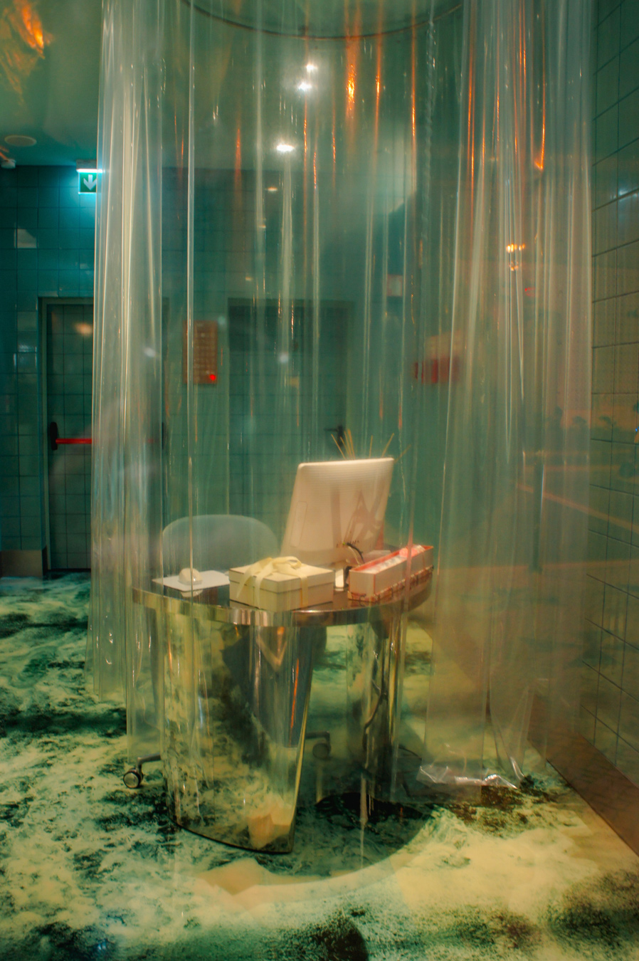Un escritorio con un ordenador y regalos se encuentra dentro de un recinto circular de cortinas de plástico transparente, en una habitación con paredes de azulejos verde azulado y un suelo que parece un remolino de agua. El escritorio es plateado y redondo, y detrás hay una silla. Fotografía de Marcia Simoes.