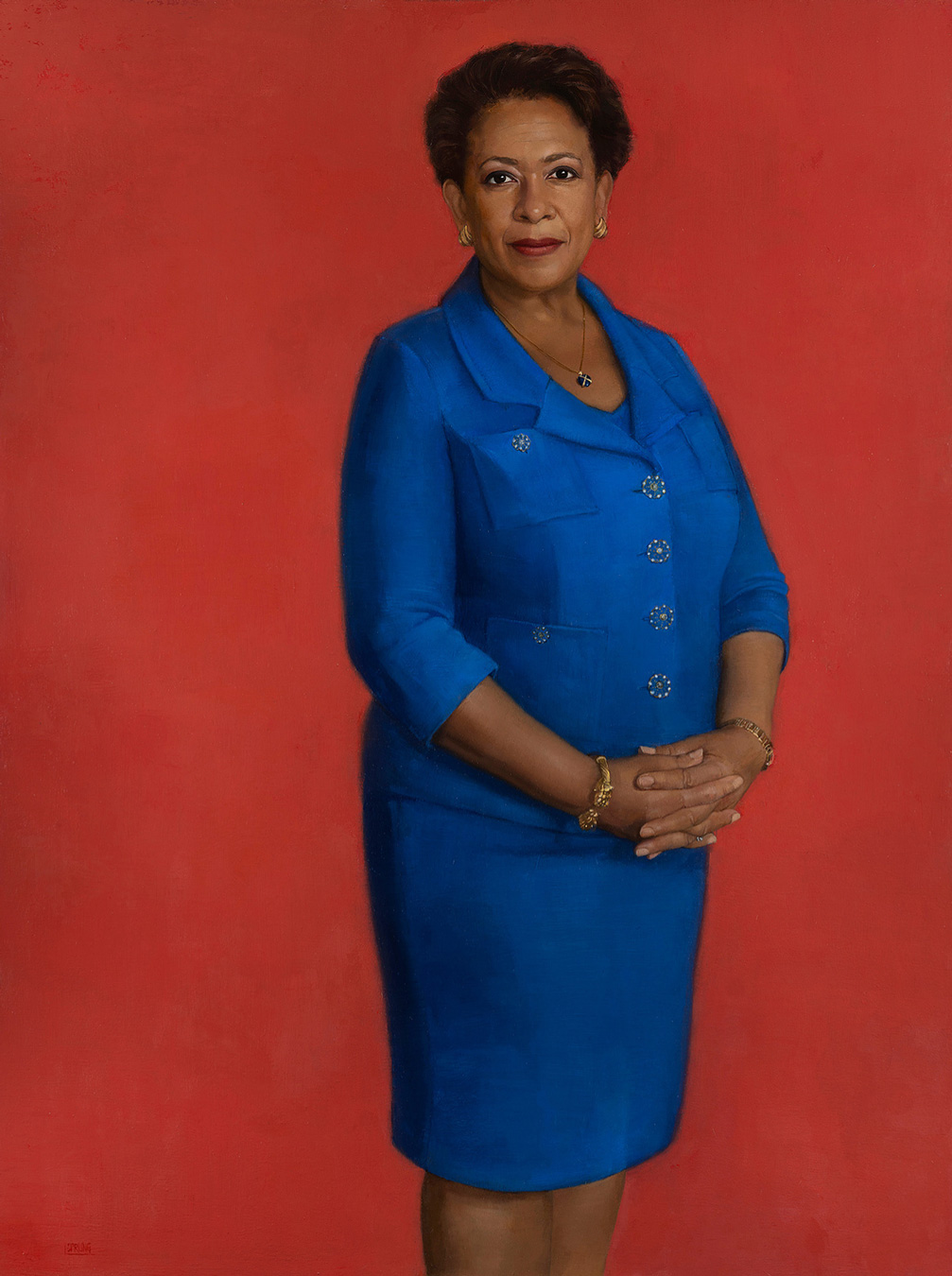 Portrait de Loretta Lynch vêtue d'un tailleur bleu sur fond rouge. Elle porte des bijoux en or et a les mains jointes.