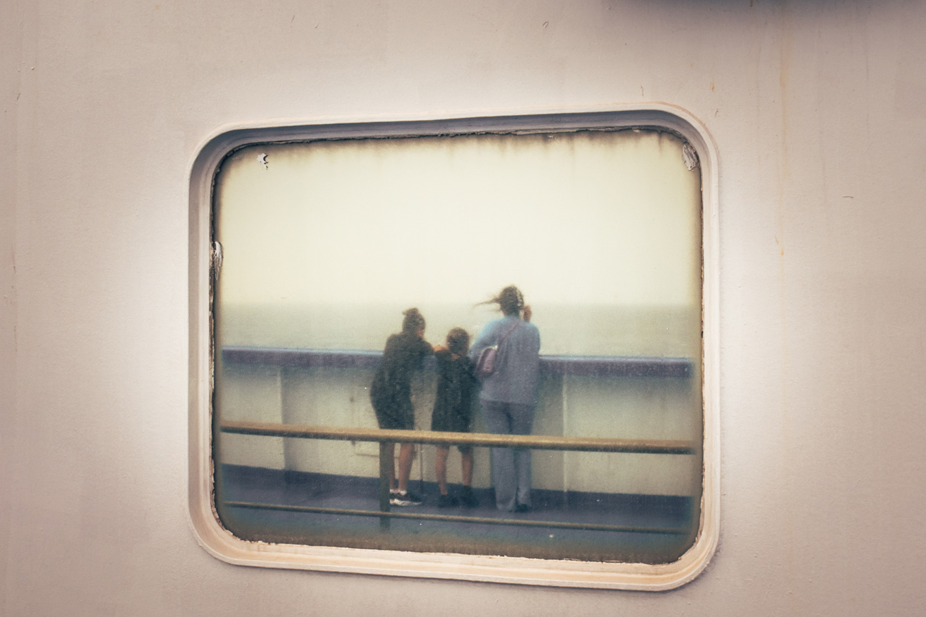 Through a weathered, rectangular window, three figures stand on a ship's deck, gazing out at the sea and sky. The window frame is off-white and shows signs of age, contrasting with the blurred, dreamlike scene beyond. Photo by Skander Khlif.