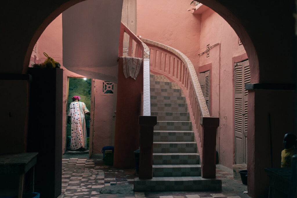 Through an archway, a tiled staircase with a pink railing leads up in a pink-walled building, while a person in a colorful robe and pink head covering walks through a doorway to the left. The scene is framed by dark foreground elements, creating a sense of depth and intrigue. Photo by Skander Khlif.