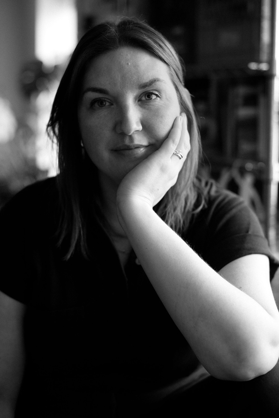 Ein Schwarz-Weiß-Porträt zeigt eine Frau mit langen Haaren, die ihr Gesicht auf ihre Hand stützt und einen Ring trägt. Sie schaut mit einem leichten Lächeln direkt in die Kamera. Foto von Sarah Bodri.