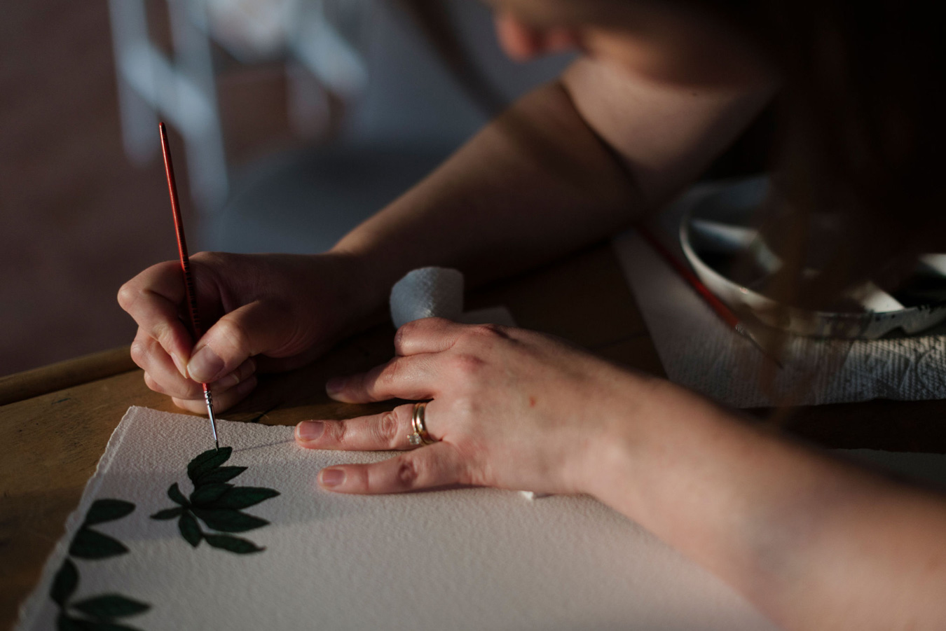 Die Künstlerin Stanzie Tooth malt mit einem dünnen roten Pinsel grüne Blätter auf weißes Papier. Die Hände der Künstlerin sind sichtbar, mit einem Ehering an einem Finger, und eine Farbpalette und ein Papiertuch liegen in der Nähe. Foto von Sarah Bodri.