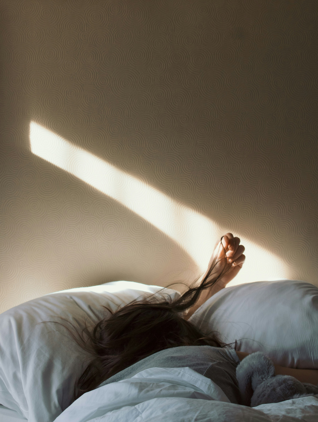Sunlight streams across a bed where a person is sleeping, head buried in pillows with an arm outstretched, illustrating the importance of restful sleep for freelancers.
