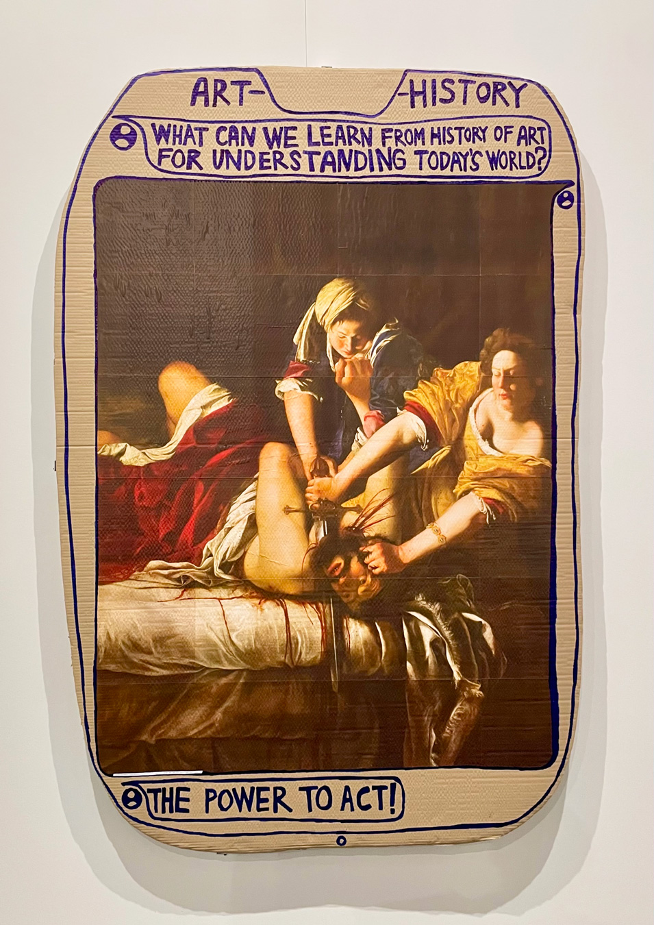 Una impresión de un cuadro que representa a Judit decapitando a Holofernes está montada sobre un trozo de cartón con una inscripción con rotulador azul que dice "ARTE-HISTORIA ¿QUÉ PODEMOS APRENDER DE LA HISTORIA DEL ARTE PARA COMPRENDER EL MUNDO DE HOY? EL PODER DE ACTUAR". Arte de Thomas Hisrchhorn