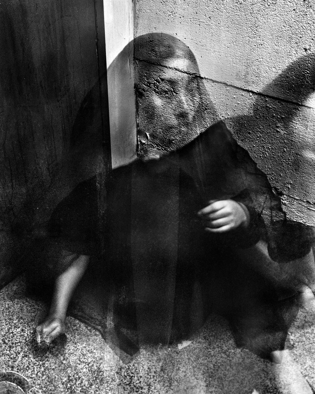 Une image granuleuse en noir et blanc montre une personne assise dans un coin, le visage masqué par un mur aux textures marquées. Ses mains sont visibles, et l'ensemble donne une impression floue et onirique.