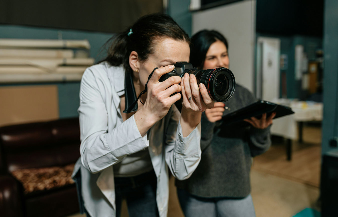 A woman in a white shirt is holding a professional camera up to her eye, taking a photo, while another woman, her assistant, stands behind her holding a tablet. The scene is indoors, within a photography studio.