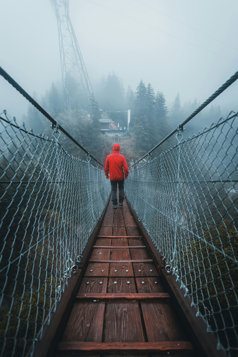 a person crossing a narrow footbridge in the fog wearing a bright red hooded coat