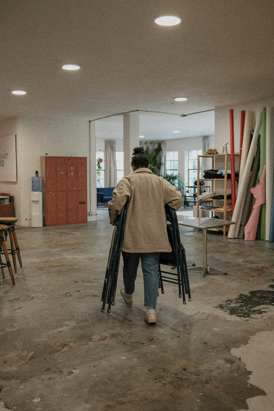 A person with a bun is seen from the back, carrying a stack of black chairs in a bright, open photography studio space. The studio features concrete floors, red lockers, and various props and furniture.