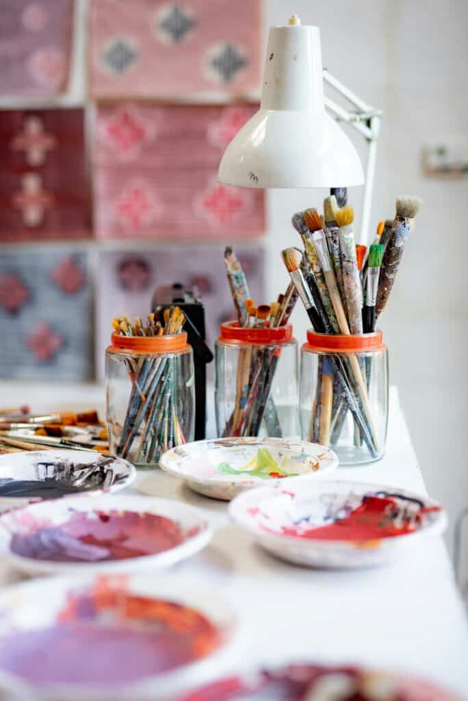 Escritorio de estudio de artista con una lámpara blanca, vasos llenos de pinceles y platos de pigmentos. Al fondo cuelgan materiales impresos a mano.