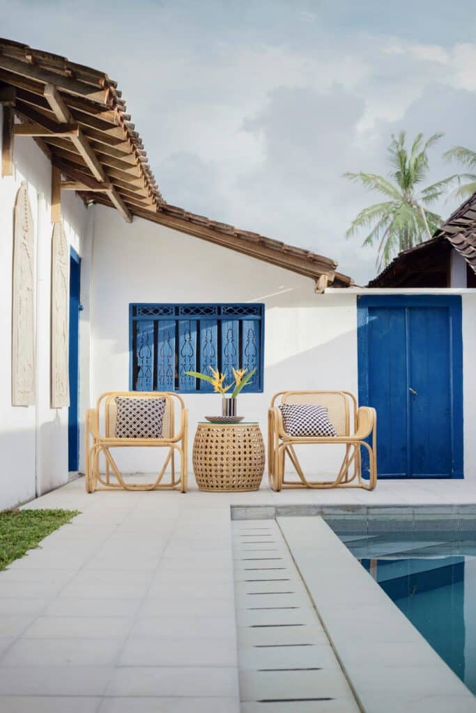 Cadeiras de bambu e mesa lateral ao lado de uma piscina externa em uma casa branca com portas e acabamentos azuis