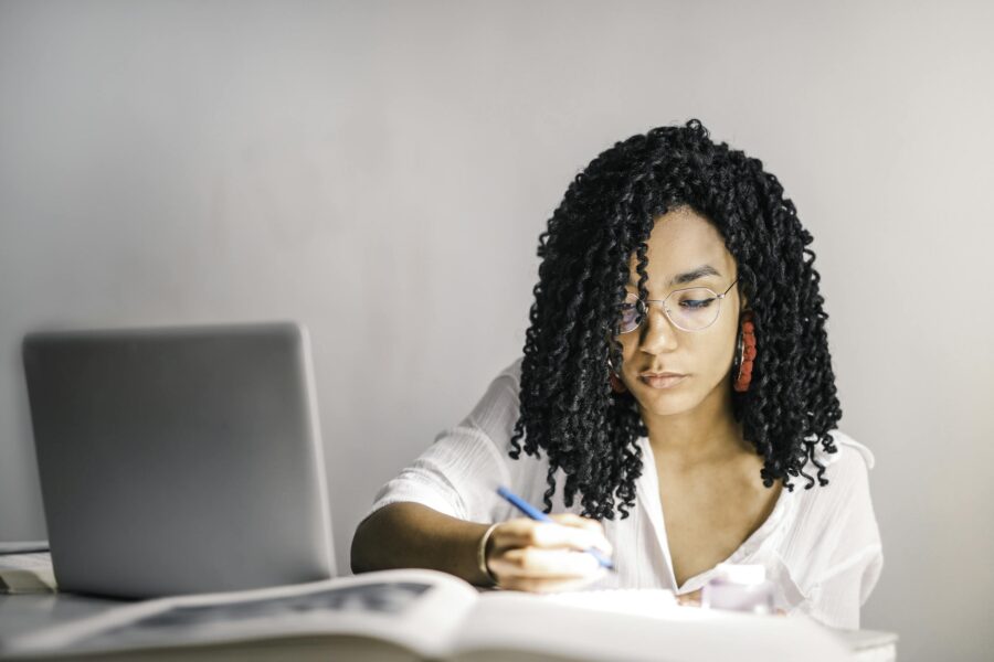 Mulher negra de camisa branca segurando uma caneta sobre um bloco de notas perto de um laptop 3765175 scaled e1706229013345