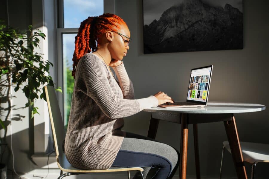 Mulher negra com cabelo vermelho sentada em uma cadeira em uma mesa redonda com um laptop sob a luz do sol e1702937763483