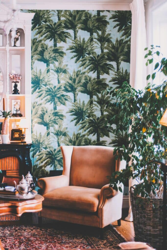 Cadeira acolchoada marrom em frente a um papel de parede com palmeiras, ao lado de um vaso de plantas e prateleiras de livros com bugigangas