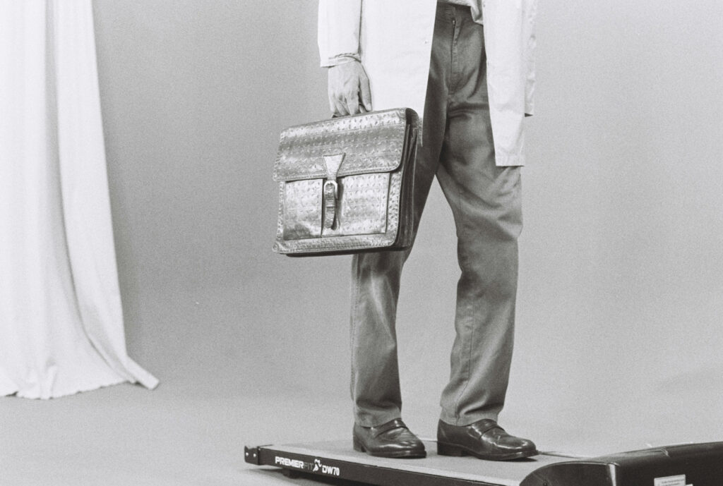 A person in a lab coat and dress shoes stands on a treadmill, holding a large, patterned briefcase. The black and white image is set against a plain background with a white curtain partially visible on the left.