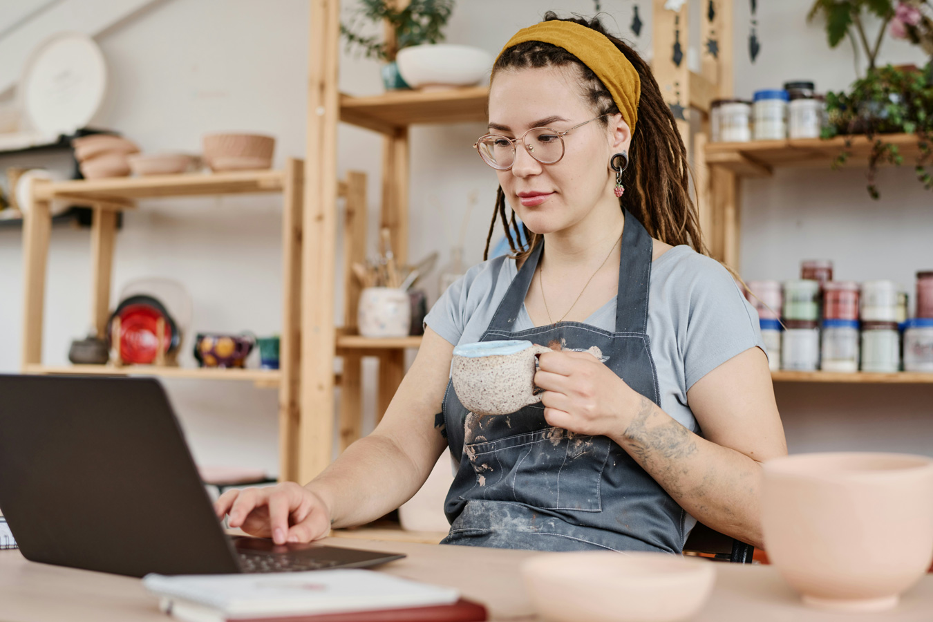 A woman with dreadlocks and glasses, wearing an apron, uses a laptop while holding a mug. Shelves with pottery and supplies are in the background.