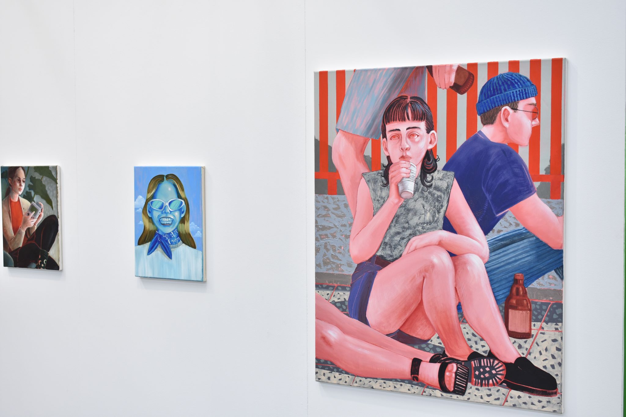 Gallery wall displaying vibrant figurative paintings, including a large scene of seated people and two portraits.