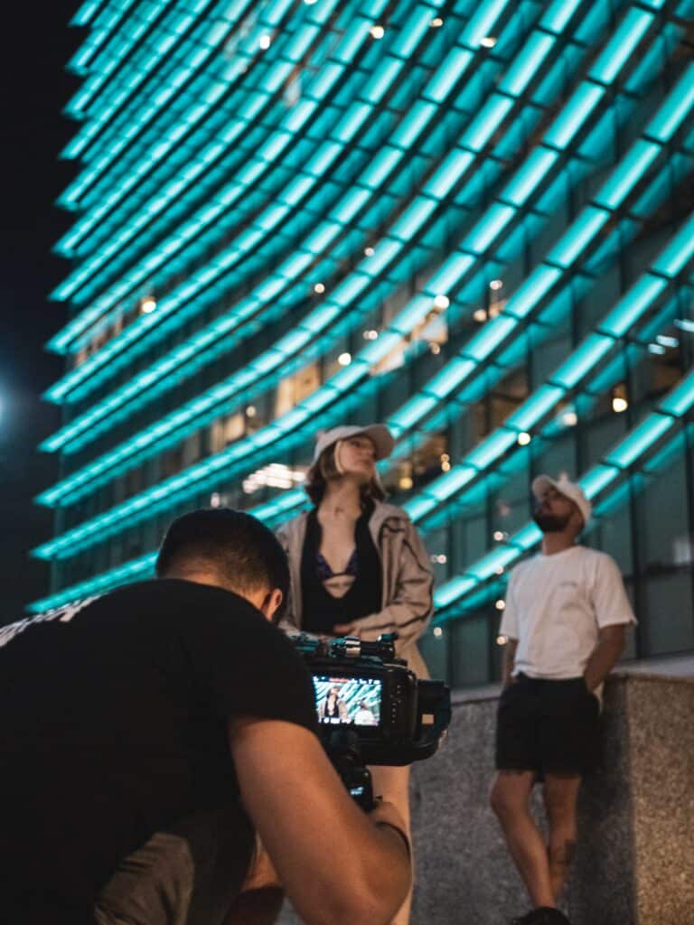 filmmaker with camera filming woman and man in front of building with aqua neon lights