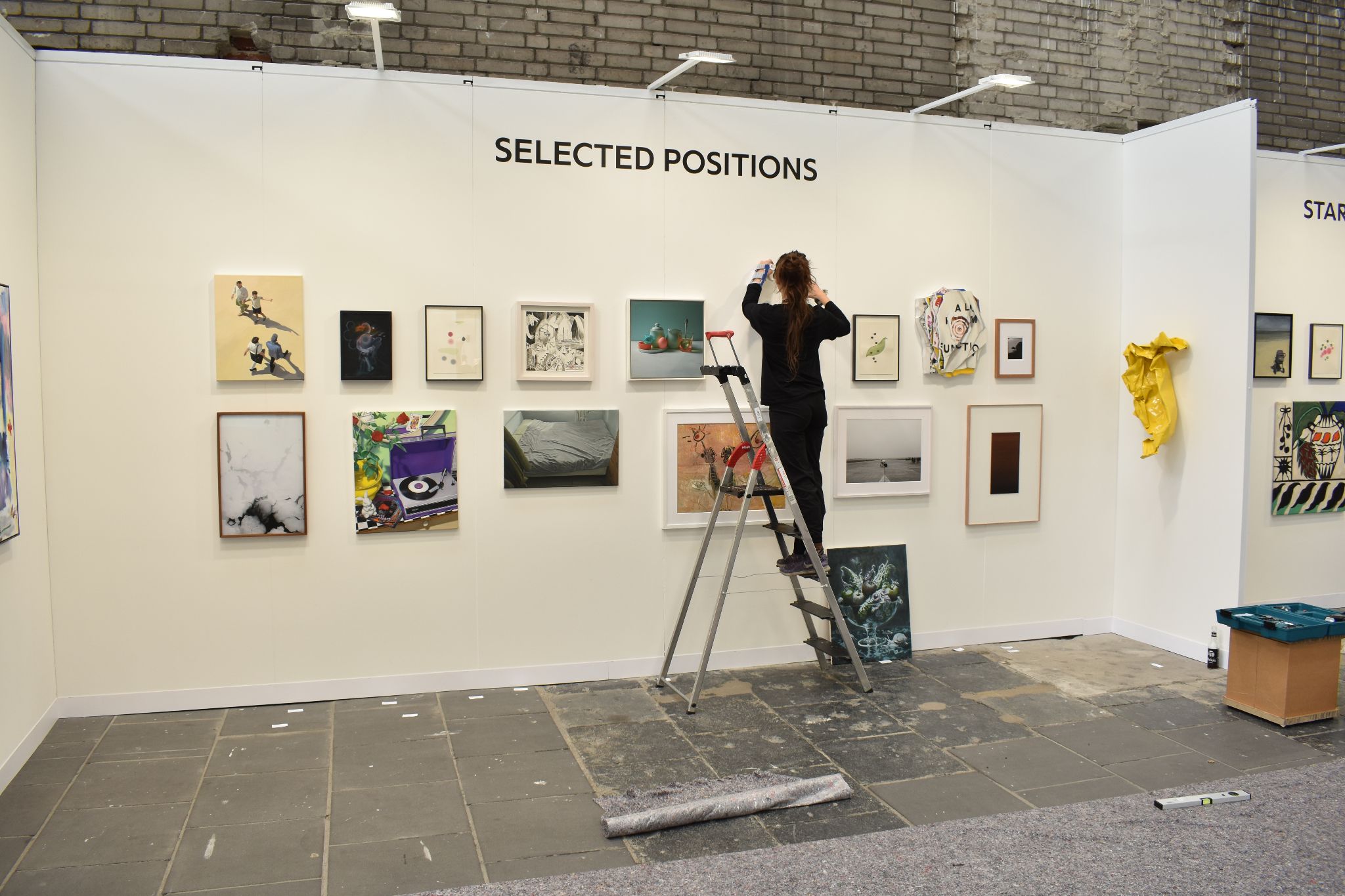 Gallery worker arranges framed artworks on an exhibition wall from a ladder.