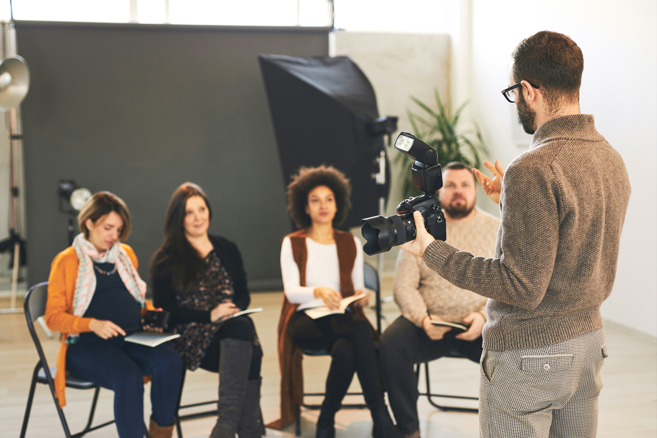 A photography instructor stands facing a group of four seated students, holding a camera with a flash attachment and gesturing with his free hand. The students, a diverse group of men and women, are seated in a studio setting with lighting equipment visible in the background.