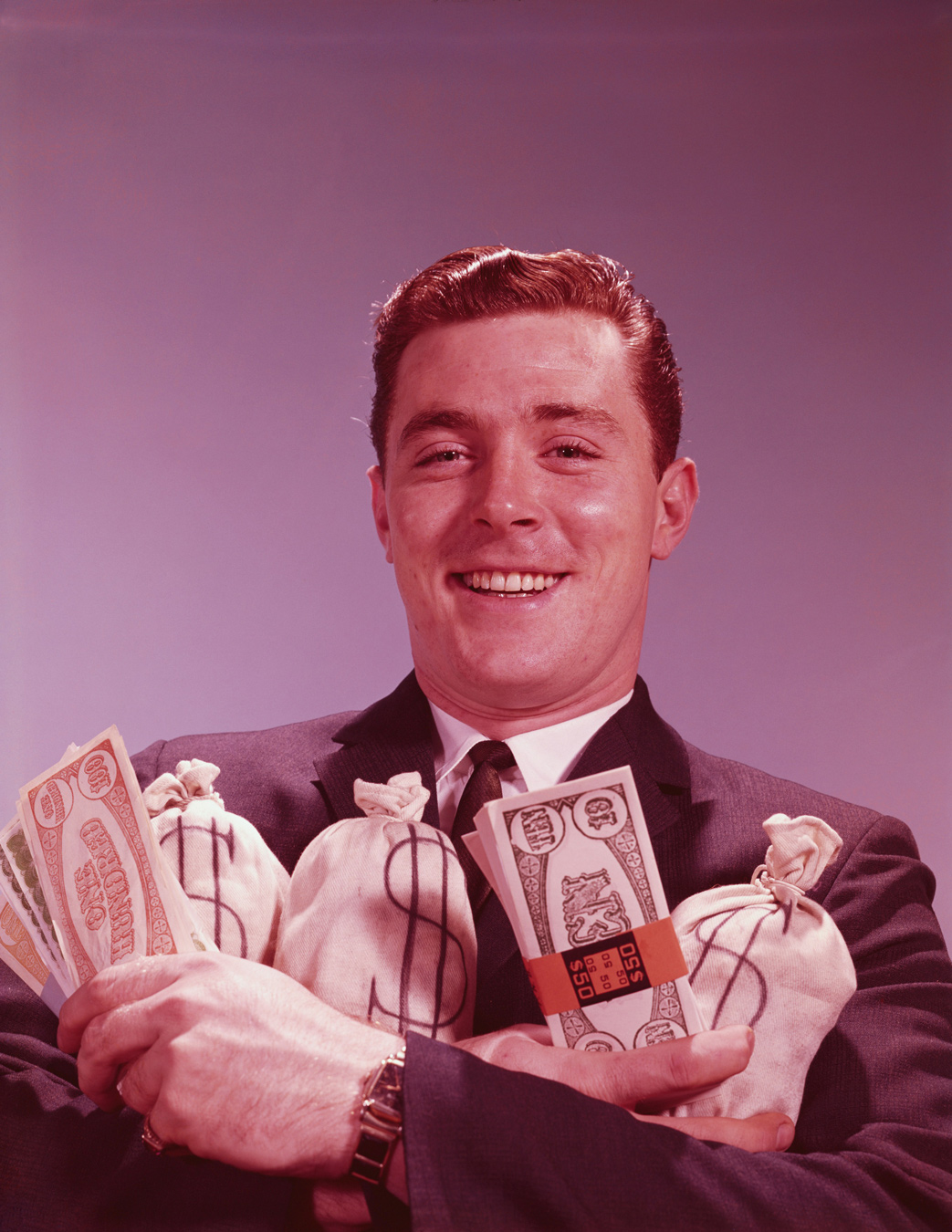 A smiling man in a suit holds stacks of cash and money bags with dollar signs on them. The man has red hair and a watch on his left wrist.