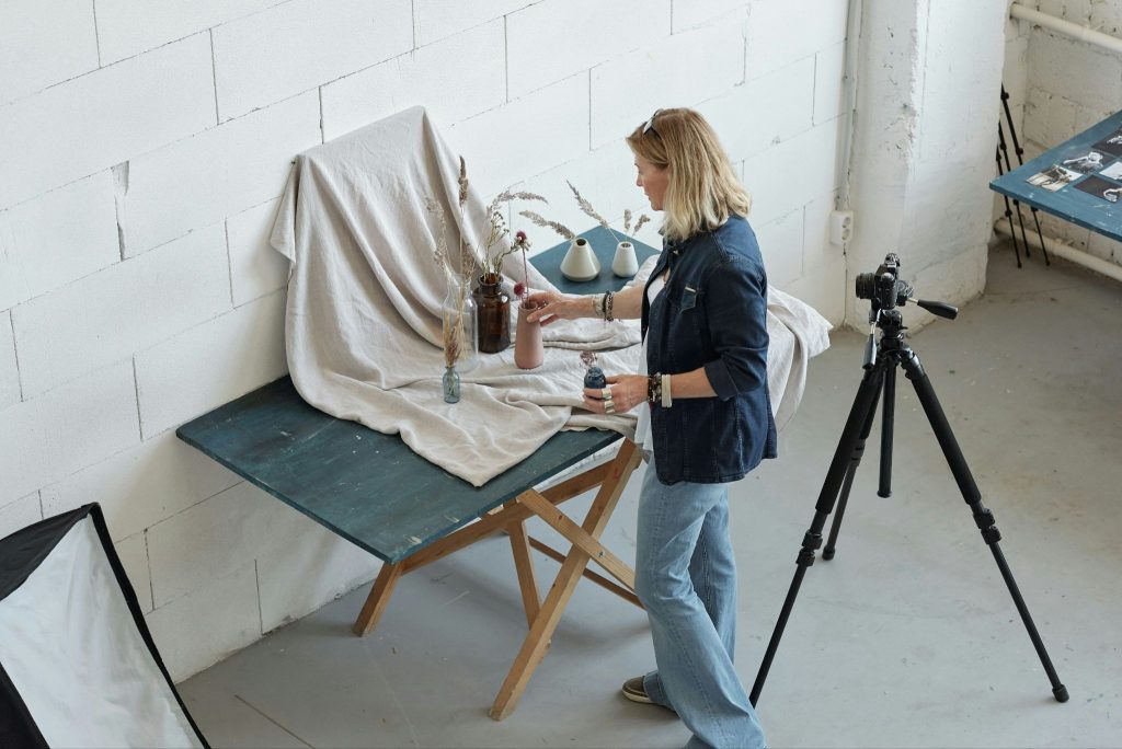 A blonde woman in a denim shirt and jeans arranges small vases with dried flowers on a draped table in a studio, with a camera on a tripod positioned nearby to capture the setup.