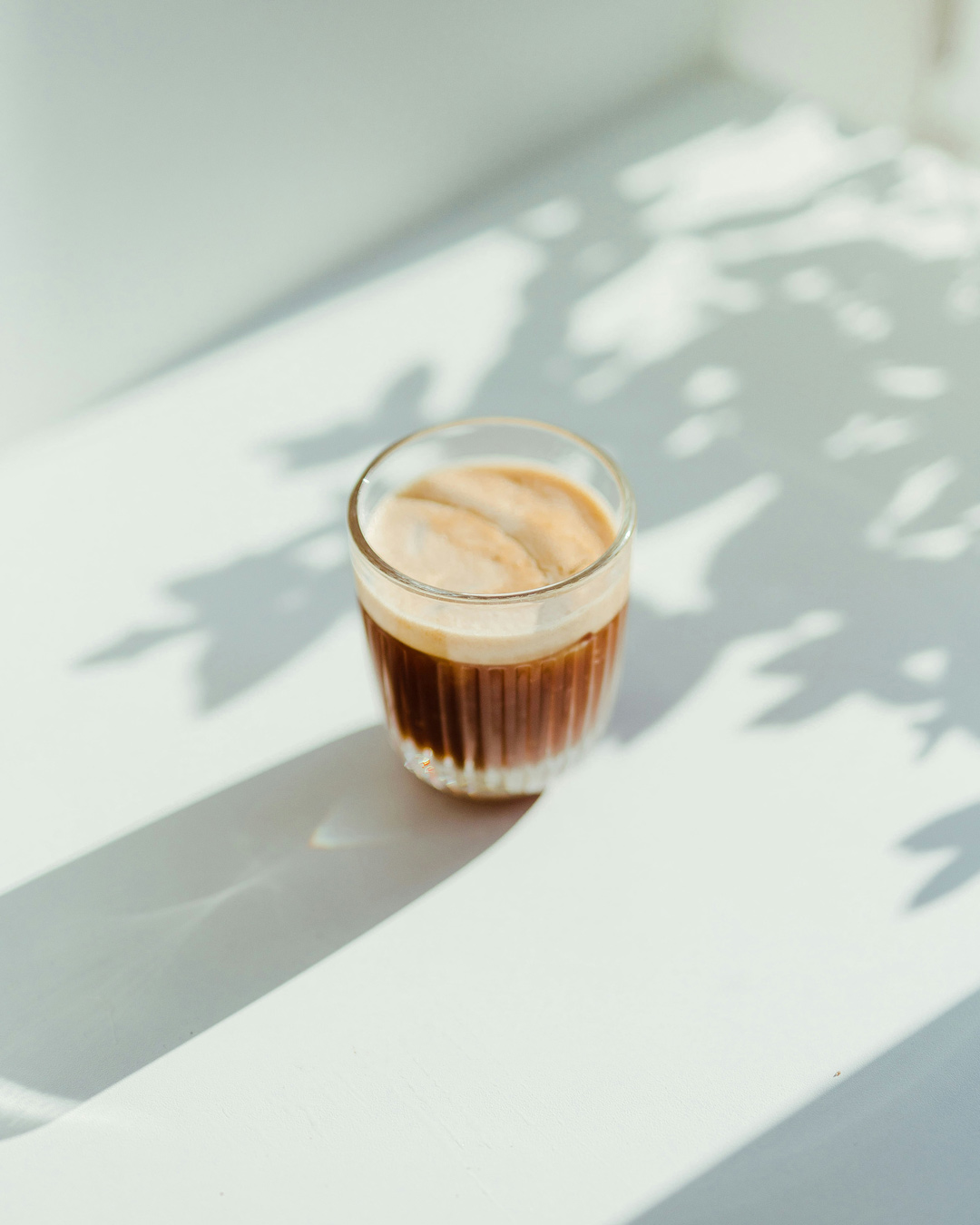 A glass of layered coffee or cacao with foam sits on a white surface, dappled with shadows, representing mindful caffeine consumption or healthier alternatives for sustained energy.
