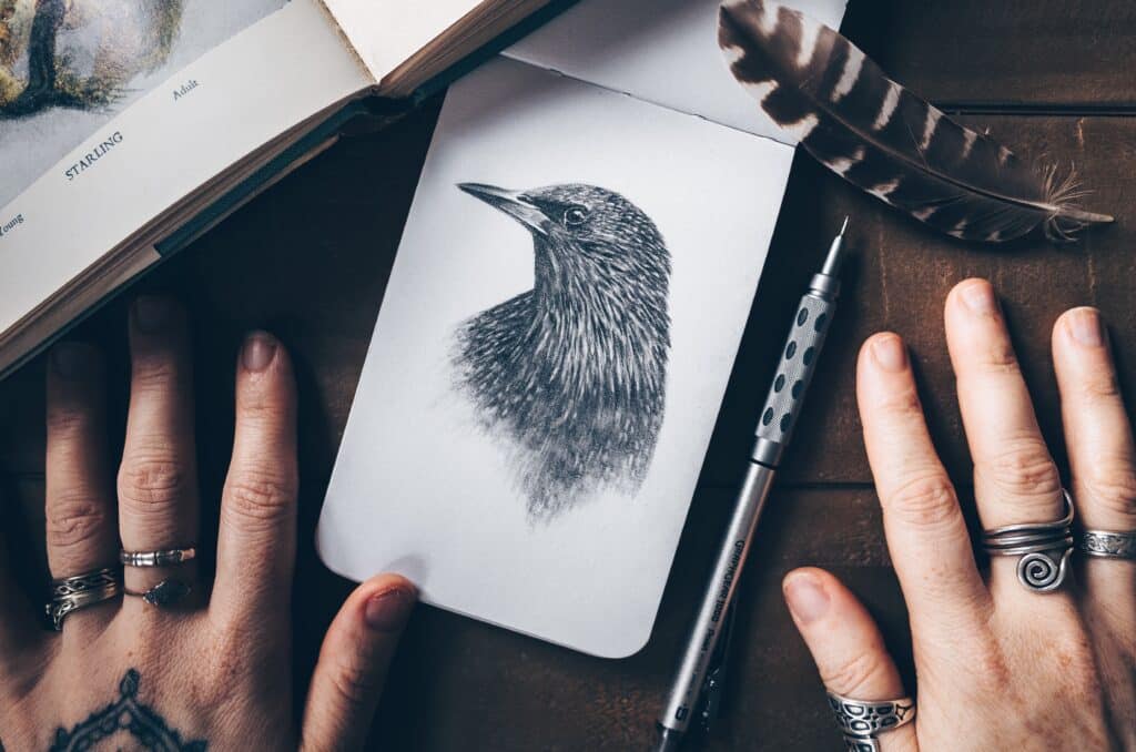 hands beside a sketch of a bird in a small unlined notebook
