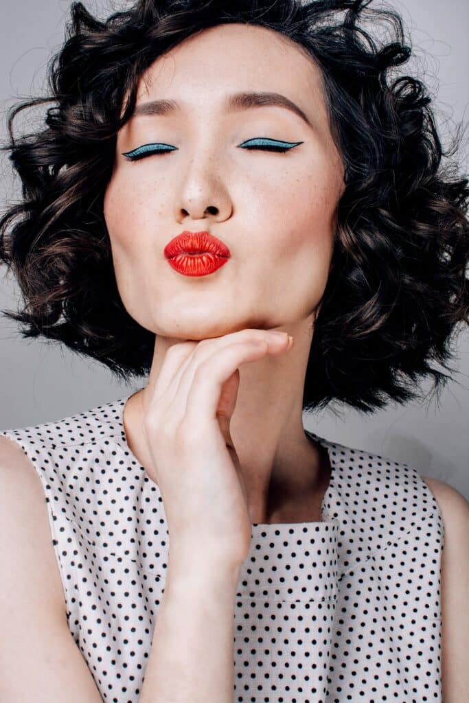 headshot of asian woman with curly hair, bright red lips pursed into a kiss, her hand curled softly under her chin
