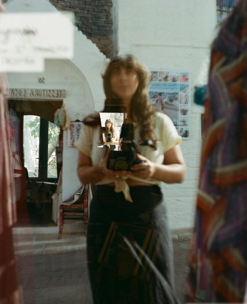 Eine Frau mit langen braunen Haaren spiegelt sich in einem Schaufenster, hält eine alte Kamera in der Hand und fotografiert ihr eigenes Spiegelbild. Der Laden hinter ihr hat eine weiße Fassade mit einem gewölbten Eingang und einem Plakat an der Wand. Selbstporträt von Sarah Bodri.
