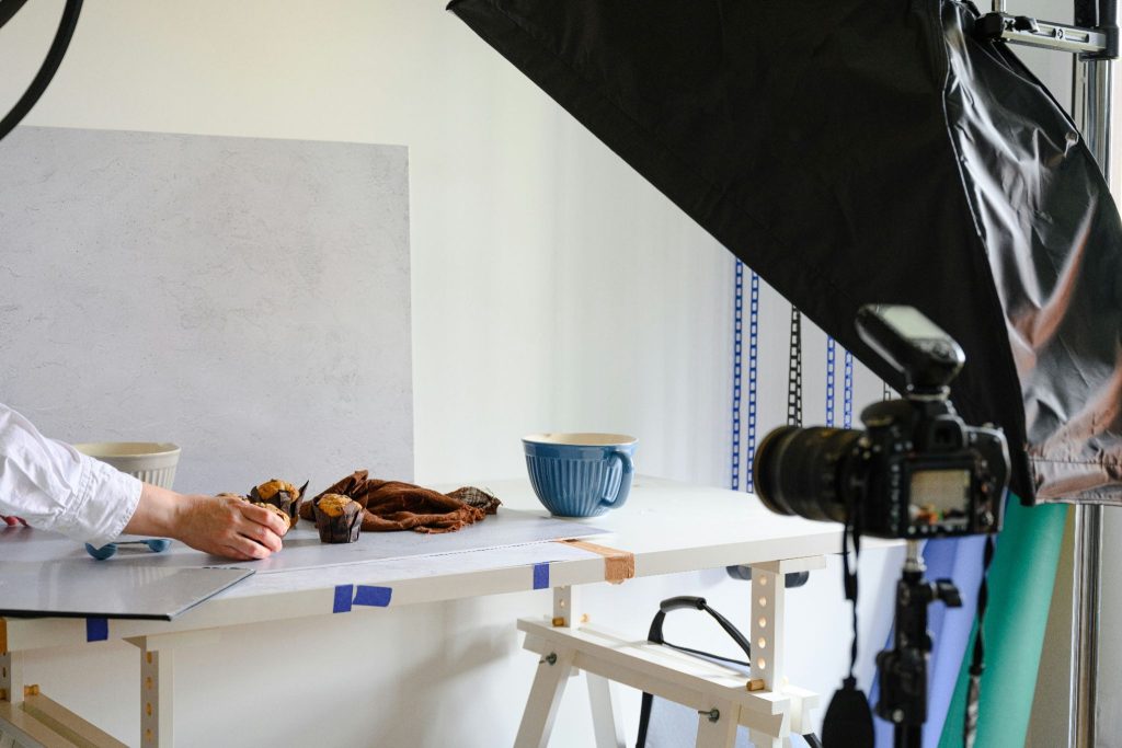 A photographer adjusts muffins on a tabletop studio setup with a blue mug and brown cloth, framed behind a DSLR camera on a tripod and a large softbox light.