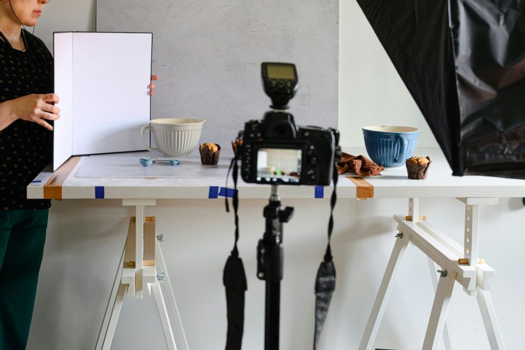 A photographer holds a white reflector board next to a food photography setup of muffins and bowls on a white table, captured from behind a DSLR camera mounted on a tripod.