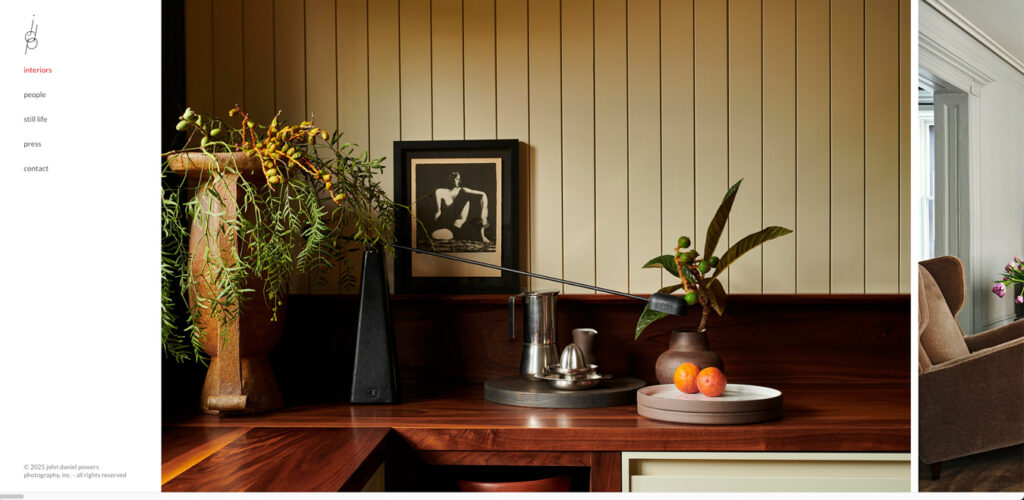 Um interior bem iluminado tem uma bancada de madeira escura cheia de itens decorativos, como um vaso com plantas, uma gravura em preto e branco emoldurada, uma cafeteira prateada e um prato com frutas. O fundo é uma parede com painéis verticais e dá pra ver uma poltrona confortável na sala ao lado.