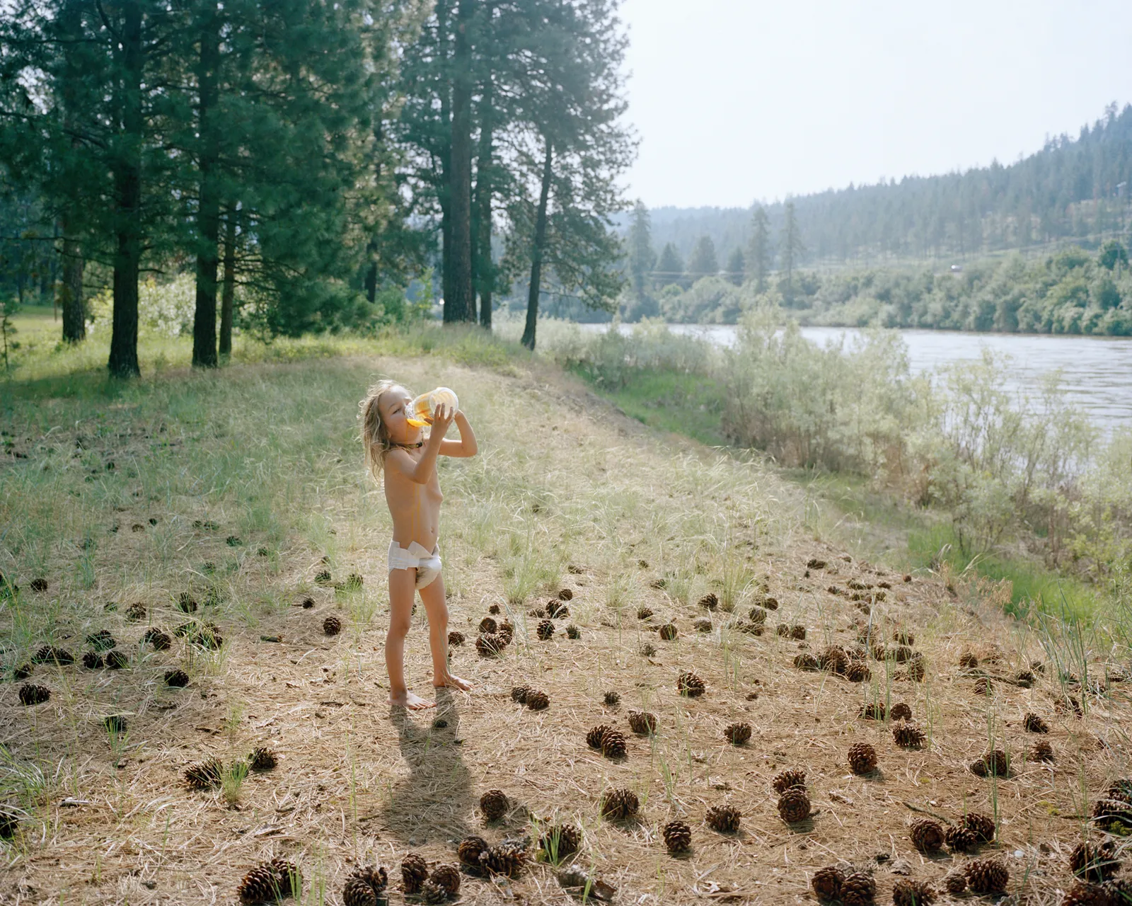 Young child drinking juice beside a river in a pine forest