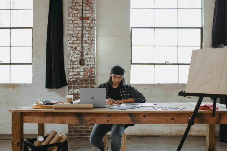 male artist sitting at a long wooden table in a studio with a laptop scaled e1703184461838