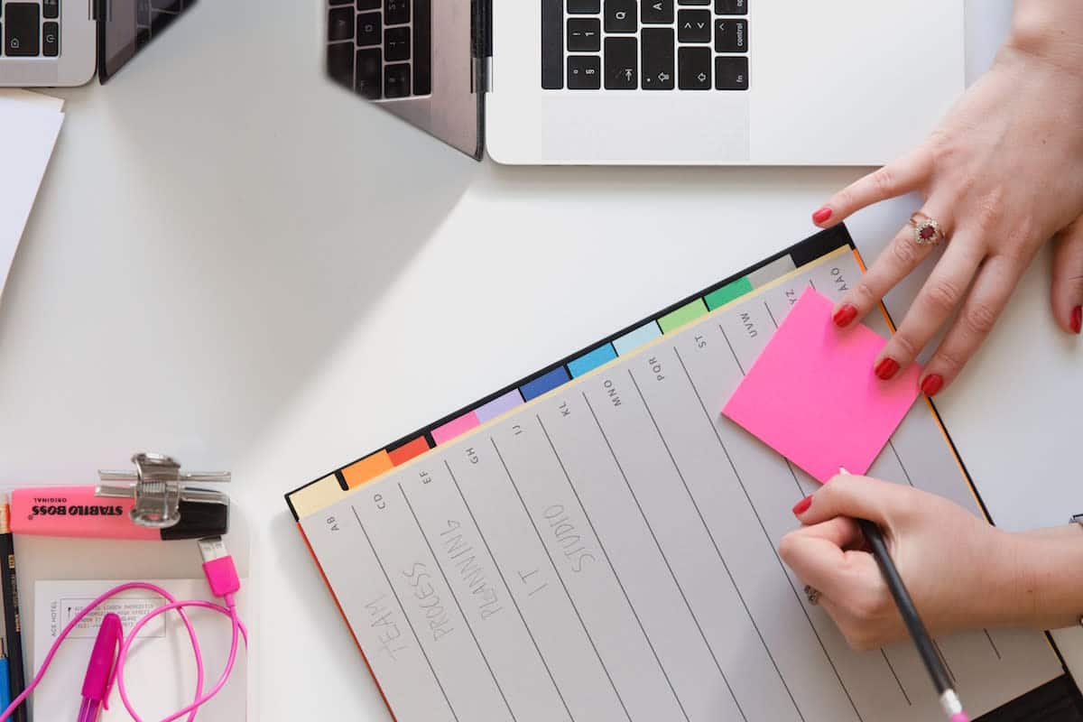 Hände, die auf einen Post-it-Zettel auf einem Planer neben einem Laptop schreiben