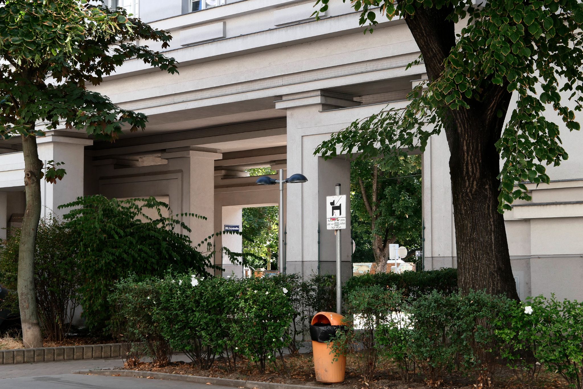 White modernist building with geometric columns, leafy trees, courtyard shrubs, and an orange trash bin