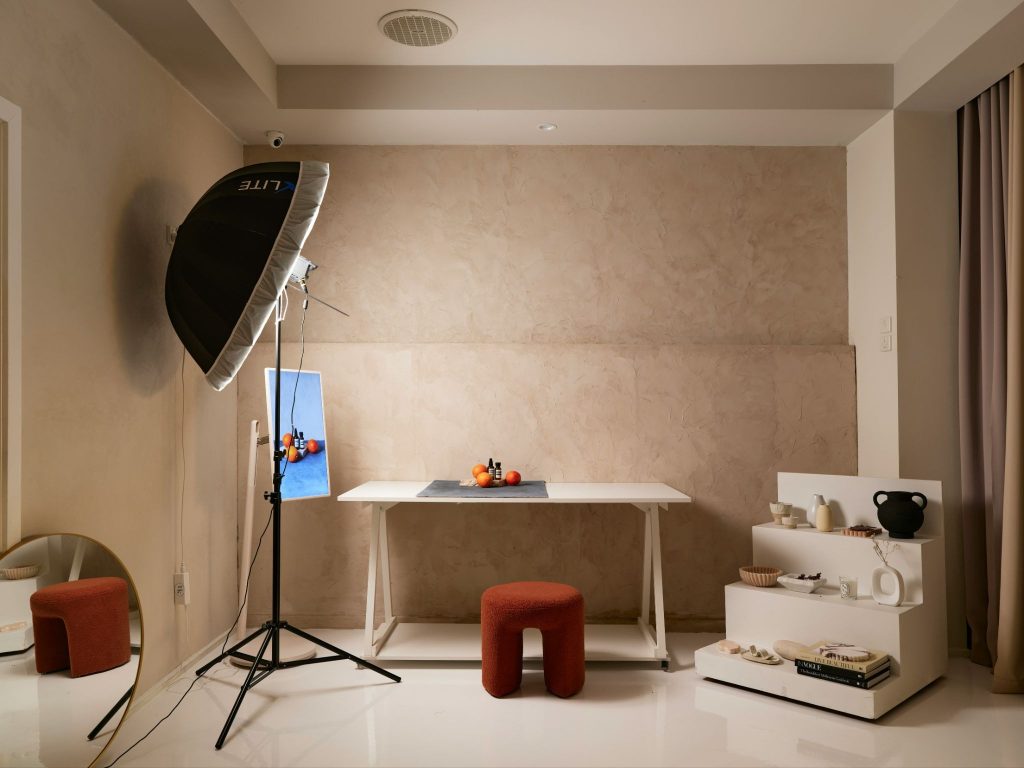 A professional photography studio setup features a white table with oranges and small bottles, illuminated by a large softbox light on a stand, with a rust-colored stool and a tiered display of decorative objects nearby against a textured beige wall.