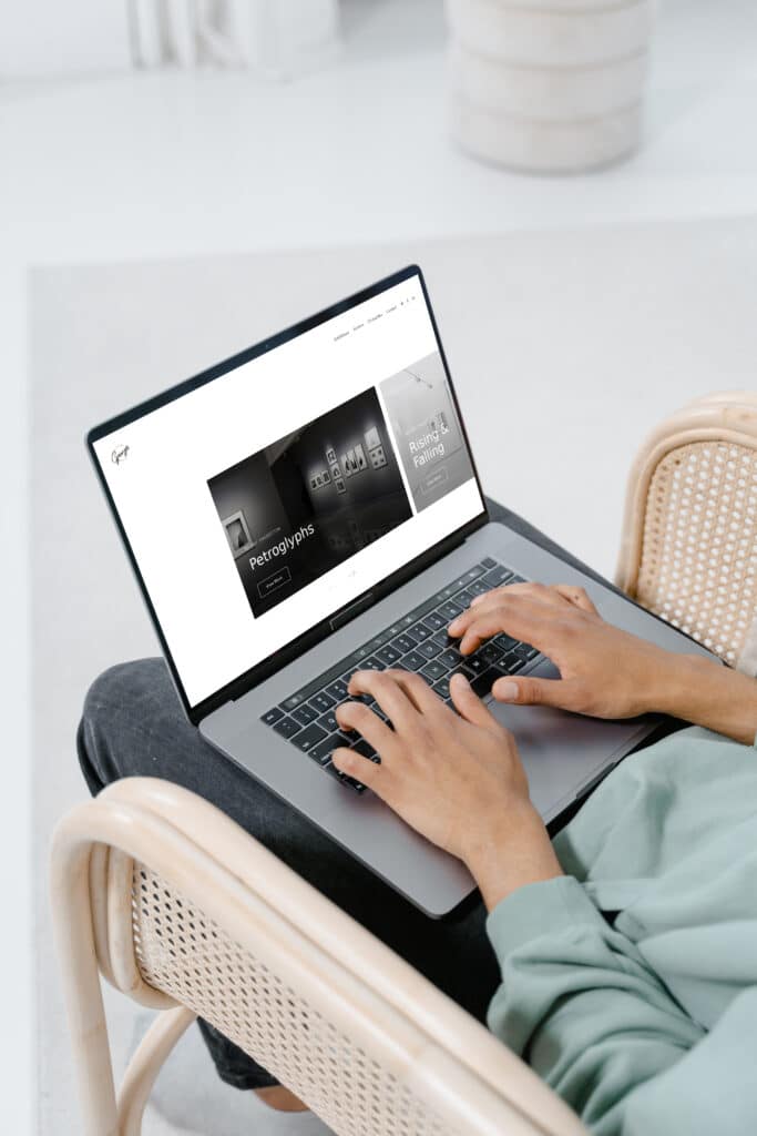 Personne assise sur une chaise, dont seules les mains et les jambes sont visibles, un ordinateur portable ouvert reposant sur ses genoux et les mains sur le clavier. L'écran affiche la page d'accueil d'un site web de format.
