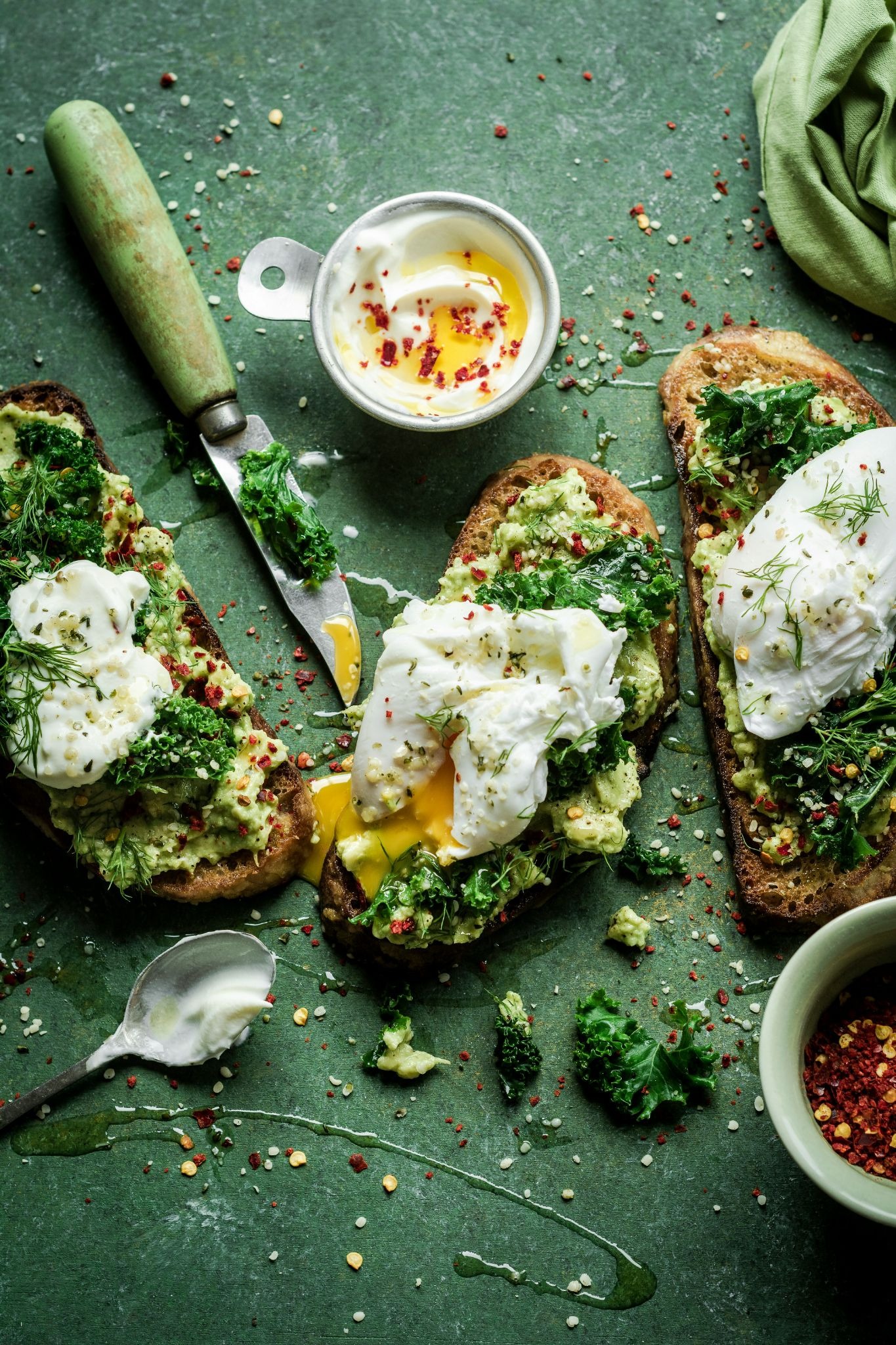 Three slices of avocado toast topped with kale and poached eggs are arranged on a green surface alongside a small bowl of yogurt, a spoon, and a knife.
