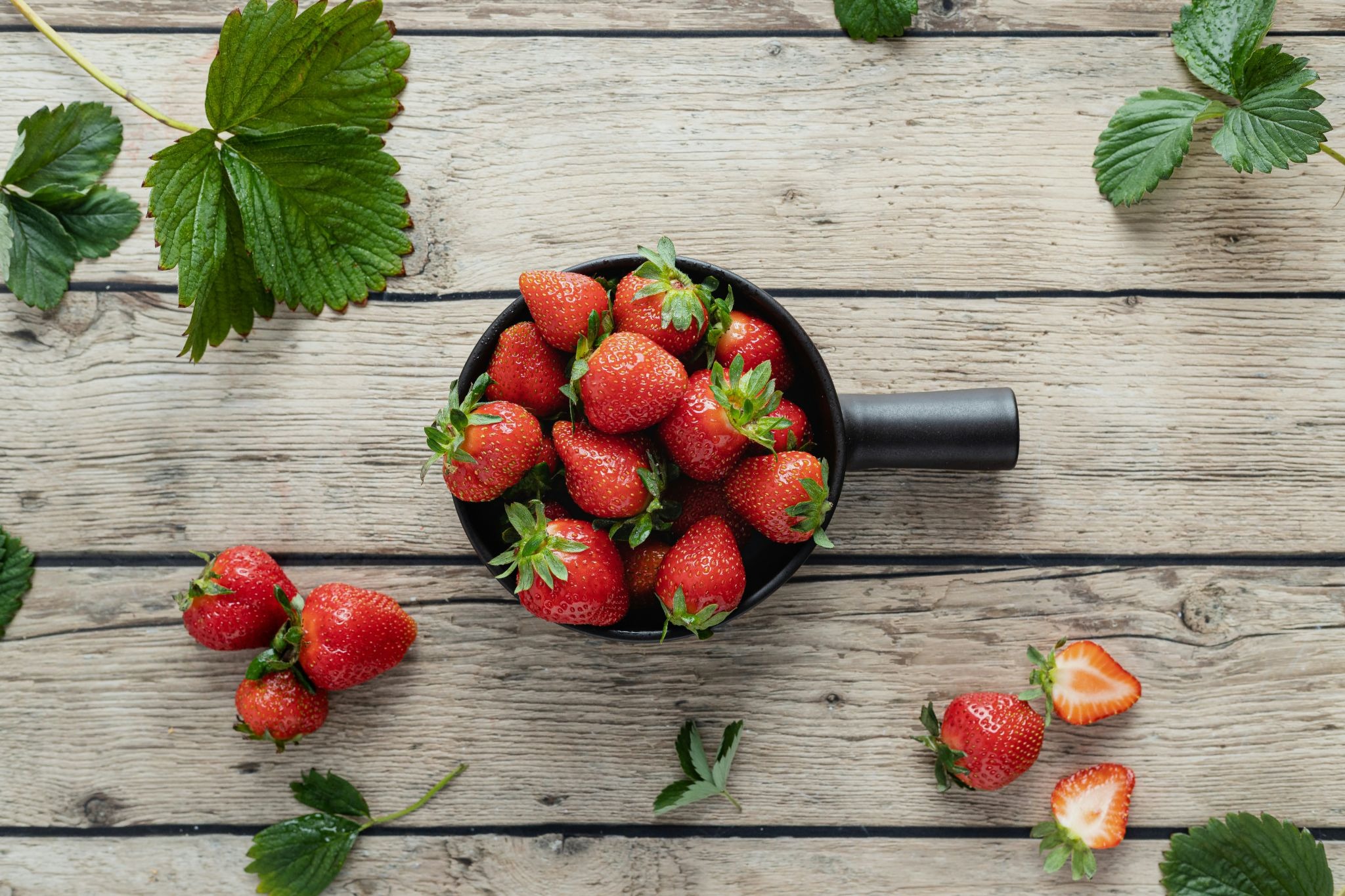 An overhead shot shows a black ceramic bowl filled with fresh, ripe strawberries, surrounded by scattered whole and halved strawberries and green leaves on a rustic wooden surface.