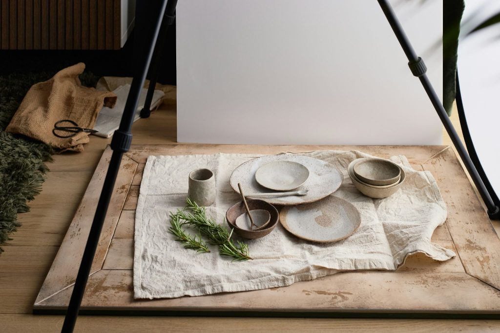 An assortment of rustic, speckled ceramic tableware, including plates, bowls, a cup, and small spoons, is arranged on a wrinkled linen cloth with a sprig of rosemary, set up for a photoshoot with a white backdrop and tripod legs framing the scene.