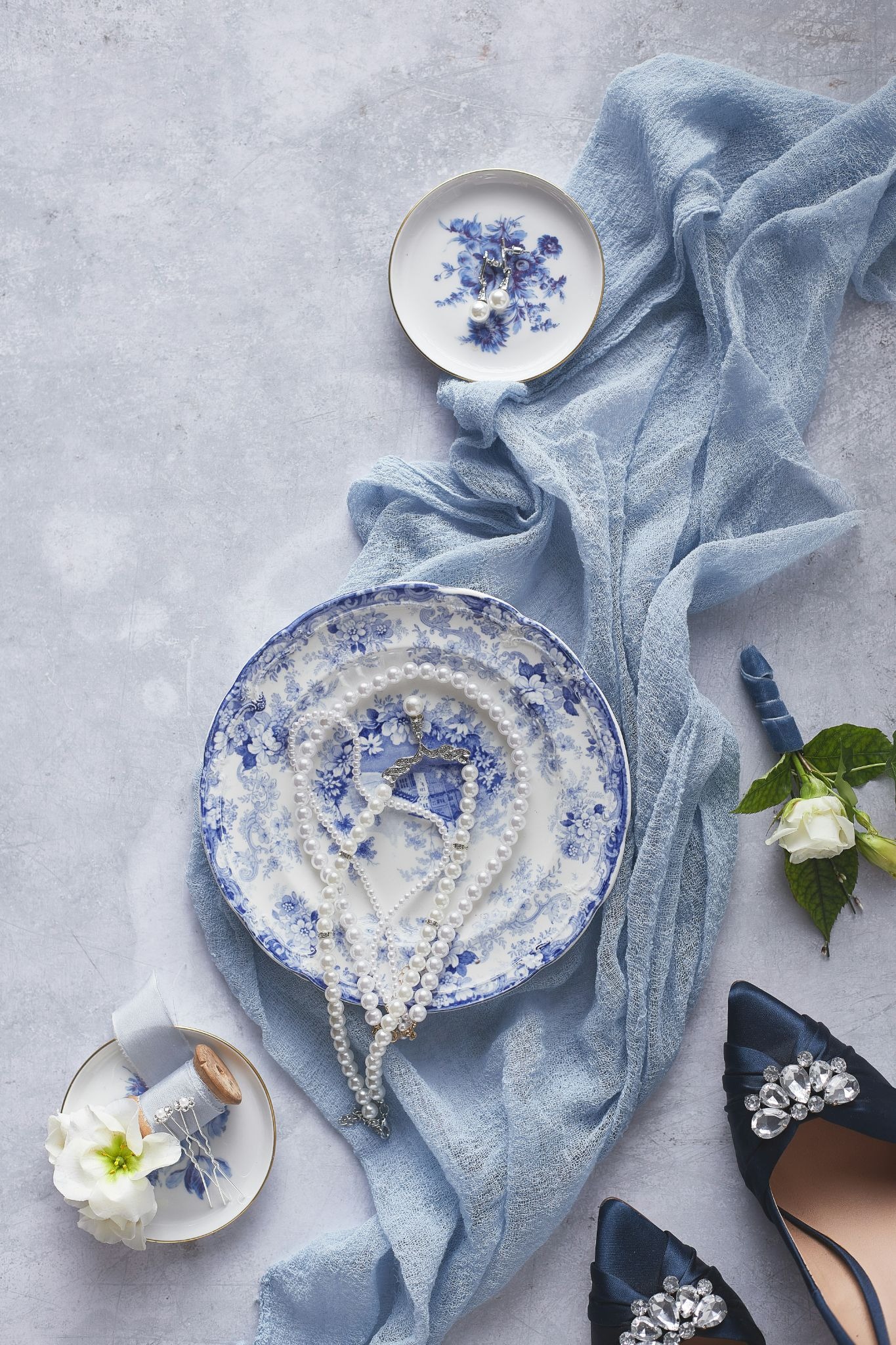 An elegant flat lay of wedding accessories features a pearl necklace on a blue and white floral plate, surrounded by a draped light blue fabric, navy blue heels with crystal embellishments, white flowers, and pearl earrings on a matching small dish.