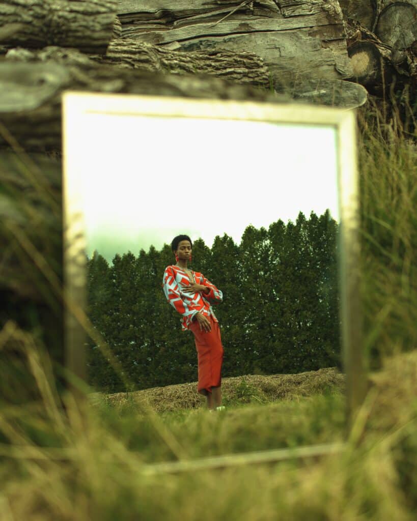 person posing in field, reflected in a mirror beside a stone wall