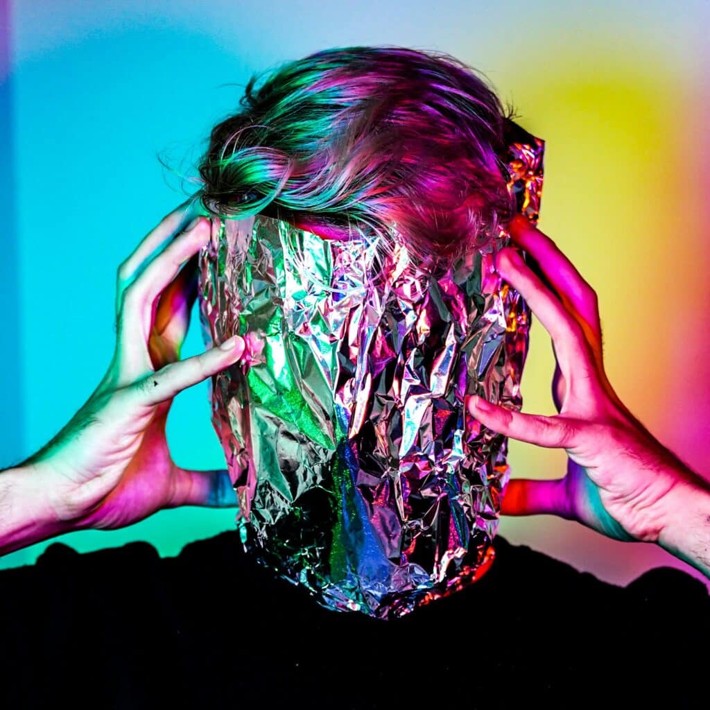 portrait of a person with aluminum foil over their face, their hands and and hair colored by neon lights