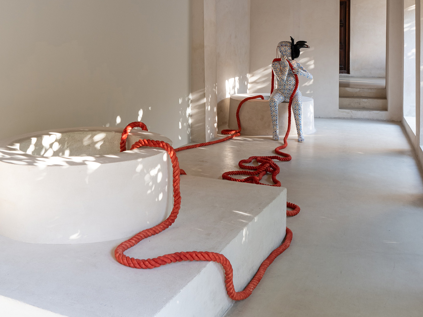 Dans une pièce lumineuse et minimaliste se trouve une sculpture aux motifs abstraits, représentant une tête d'oiseau noire, posée sur un socle blanc. Une épaisse corde rouge s'enroule autour d'éléments architecturaux blancs et rejoint la sculpture. La lumière du soleil pénètre à flots par les fenêtres.