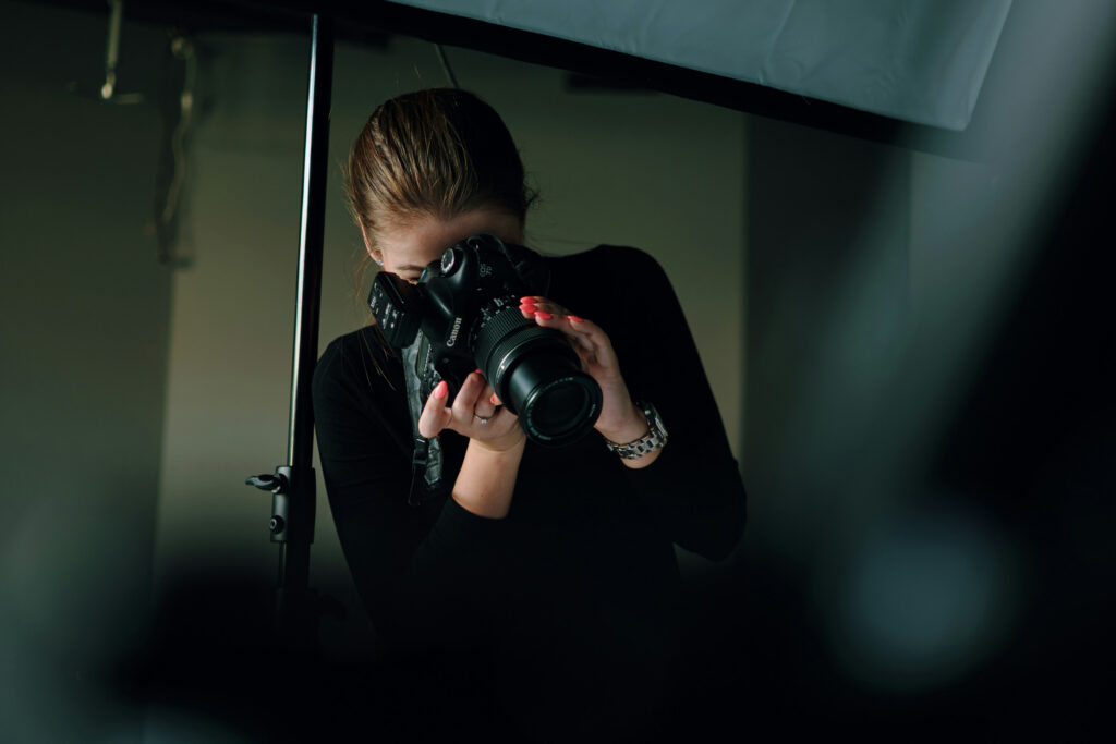 A woman holds a Canon EOS 7D camera with a zoom lens, aiming it forward. She has red nail polish and wears a watch. A light stand is visible to the left.